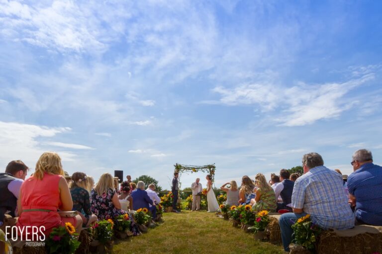 Outdoor wedding ceremony in a field with guests seated on either side facing a decorated arch with sunflowers.