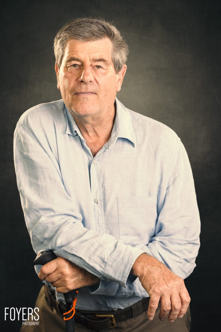 A middle-aged man in a light blue shirt stands against a dark background, looking at the camera with a neutral expression.