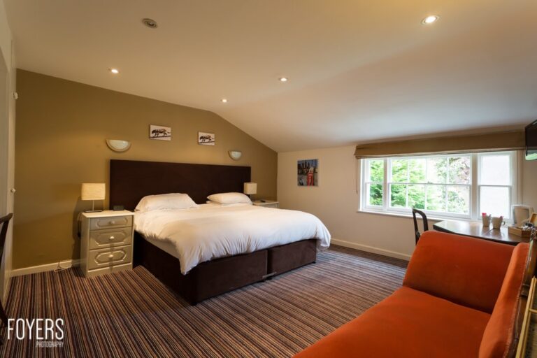 A spacious bedroom with a dark wooden bed, white bedding, olive walls, striped carpet, orange armchair, and large windows overlooking greenery.