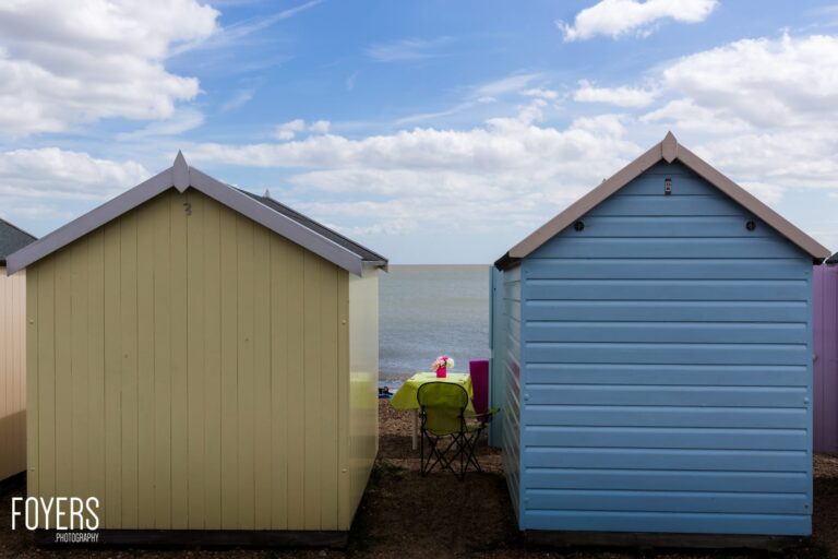 Two colourful beach huts frame a person sitting in a chair between them, facing the sea on a sunny day.