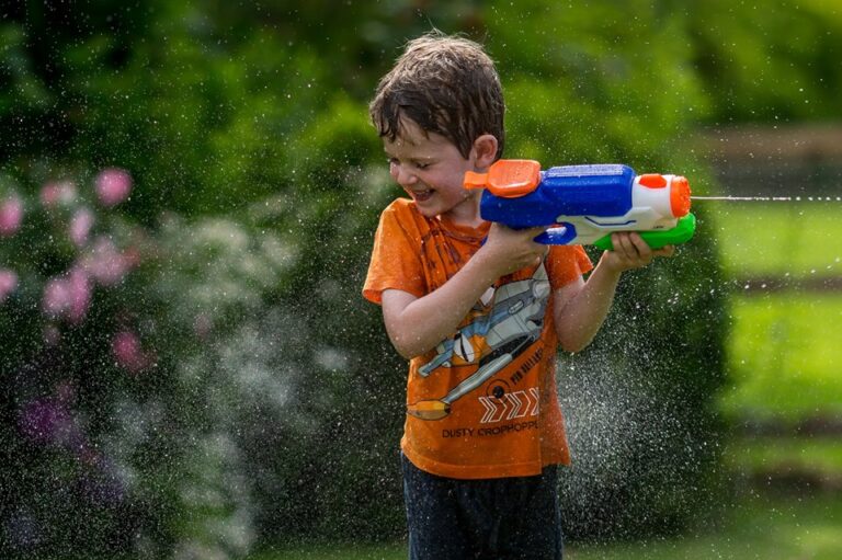A young boy in an orange shirt holds a toy water gun, spraying water outdoors on a sunny day with green foliage behind him.