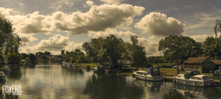 Moored boats on a calm river with trees and buildings along the banks under a cloudy sky.
