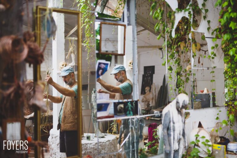 Two workers in caps and aprons work in a narrow courtyard surrounded by climbing ivy, mirrors and sculptural pieces.