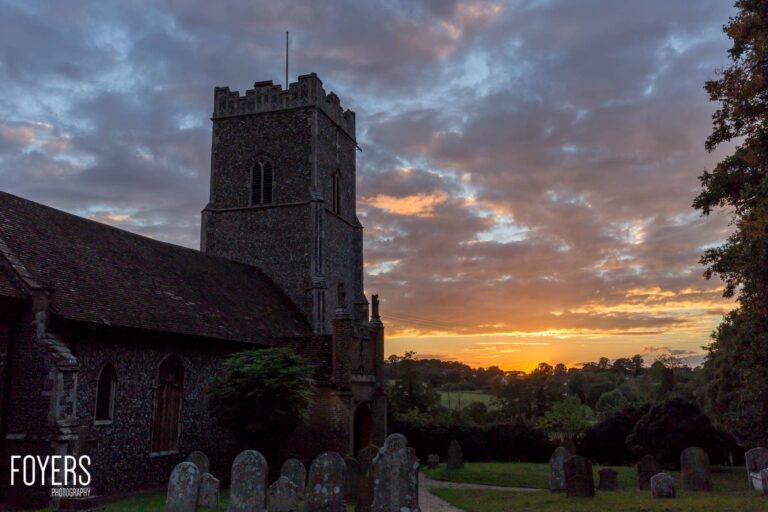 A stone church with a crenellated tower stands at sunset, surrounded by gravestones in the churchyard.