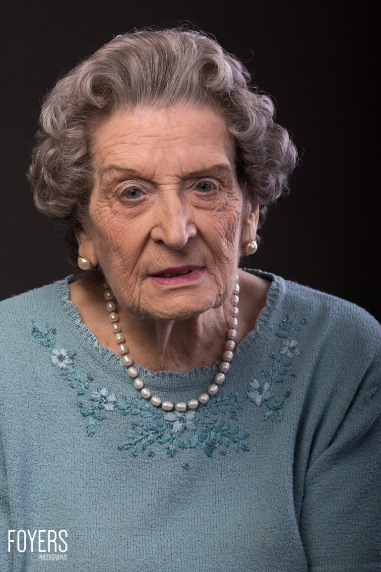 Elderly woman with grey wavy hair wearing a pale blue embroidered sweater and pearl necklace and earrings against a dark background.