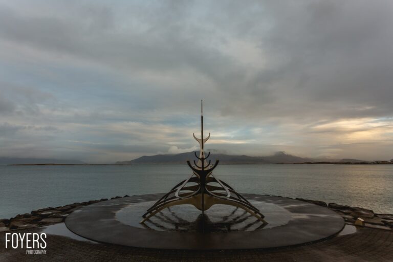 A bronze Viking ship sculpture stands in shallow water on a beach, with mountains visible across the bay under an overcast sky.