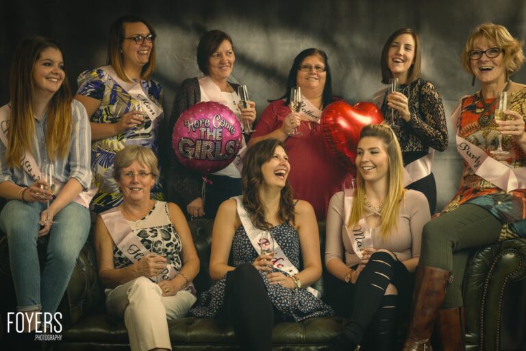 A group of women gathered for a celebration, holding champagne glasses and a Here Come the Girls balloon, smiling at the camera.