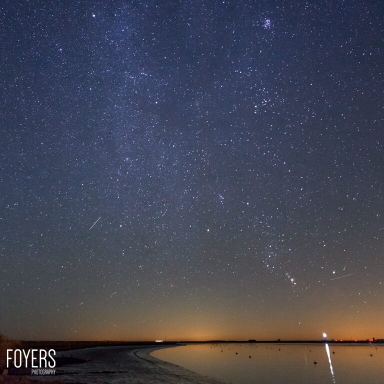 Starry night sky above a calm, reflective body of water at dusk, with several meteors streaking across the sky.