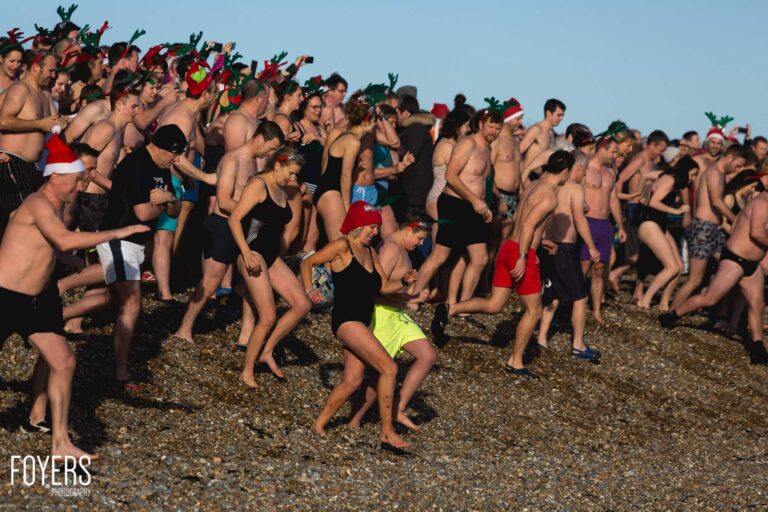 Large group of people in swimming attire and festive headwear gathered on a pebbly beach on a clear day.