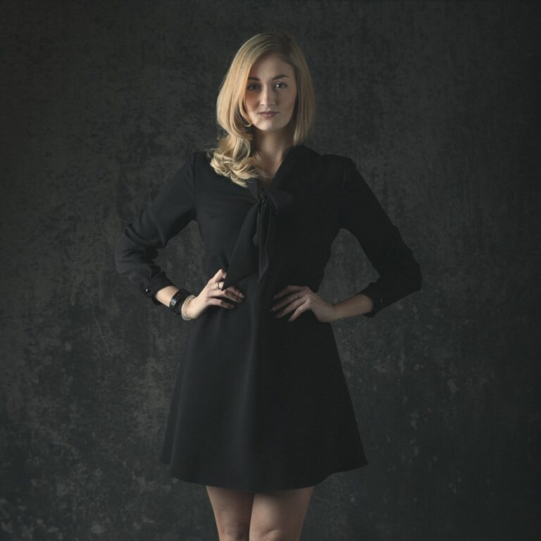 A woman with shoulder-length blonde hair wearing a black coat dress stands against a dark textured background, looking toward the camera.