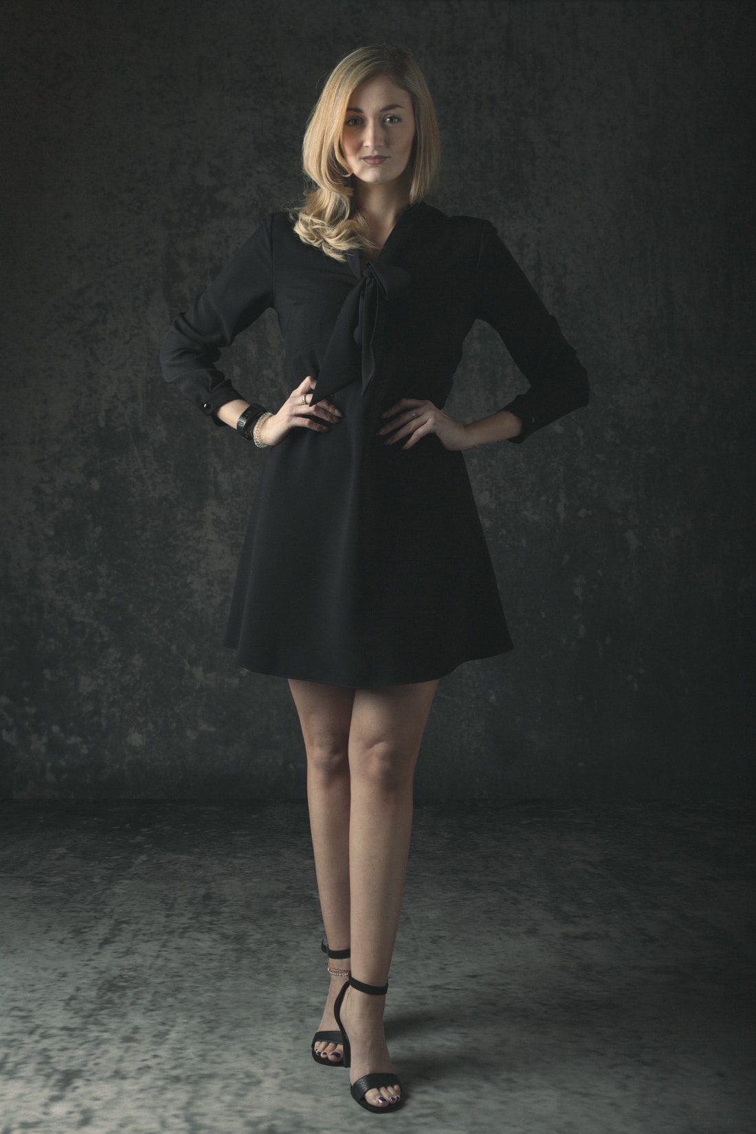 A woman with blonde hair wearing a black dress and black sandals stands against a dark textured background.