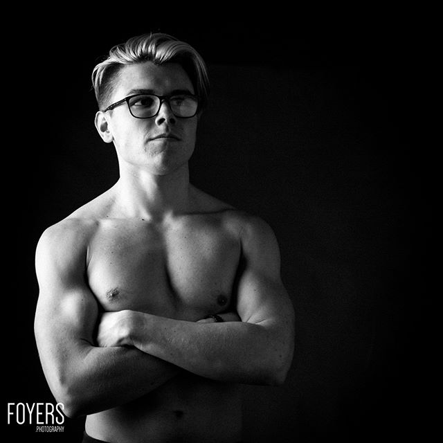 A shirtless young man wearing glasses stands against a black background with arms crossed, looking upward. Black and white portrait.