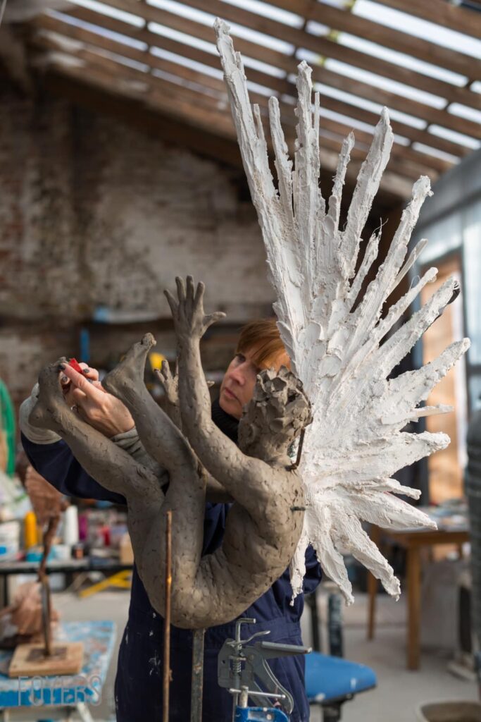 An artist holds a large white plaster sculpture of a winged figure in an industrial studio space.