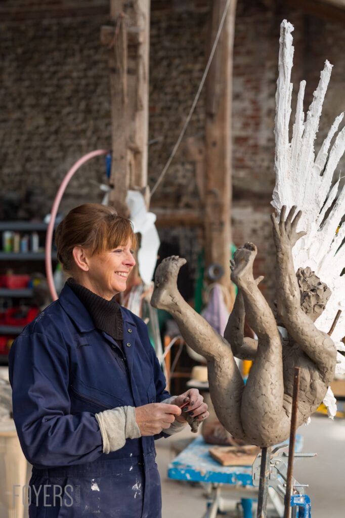 A woman in a navy work jacket smiles while examining a large clay sculpture in an art studio.