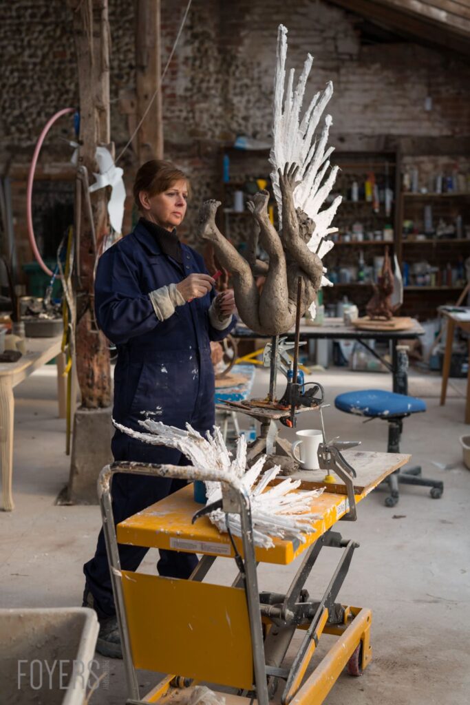 Artist in navy workwear painting white wings onto a clay sculptured figure in an industrial studio workshop.
