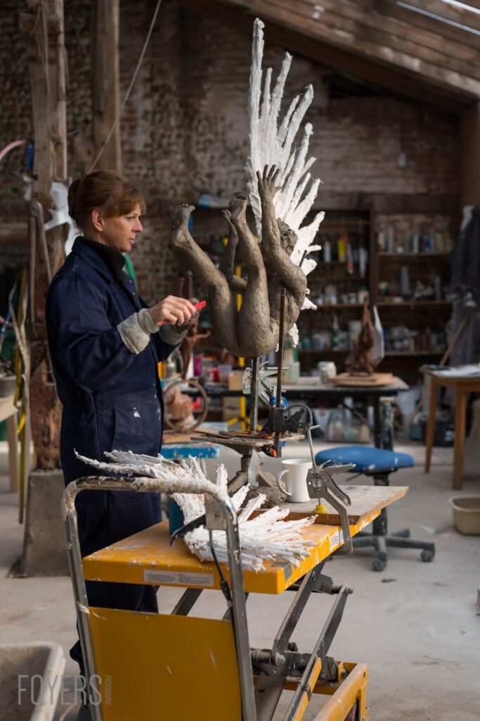 A sculptor in a navy boilersuit works on a large clay figure with white-painted wings in a studio workshop.