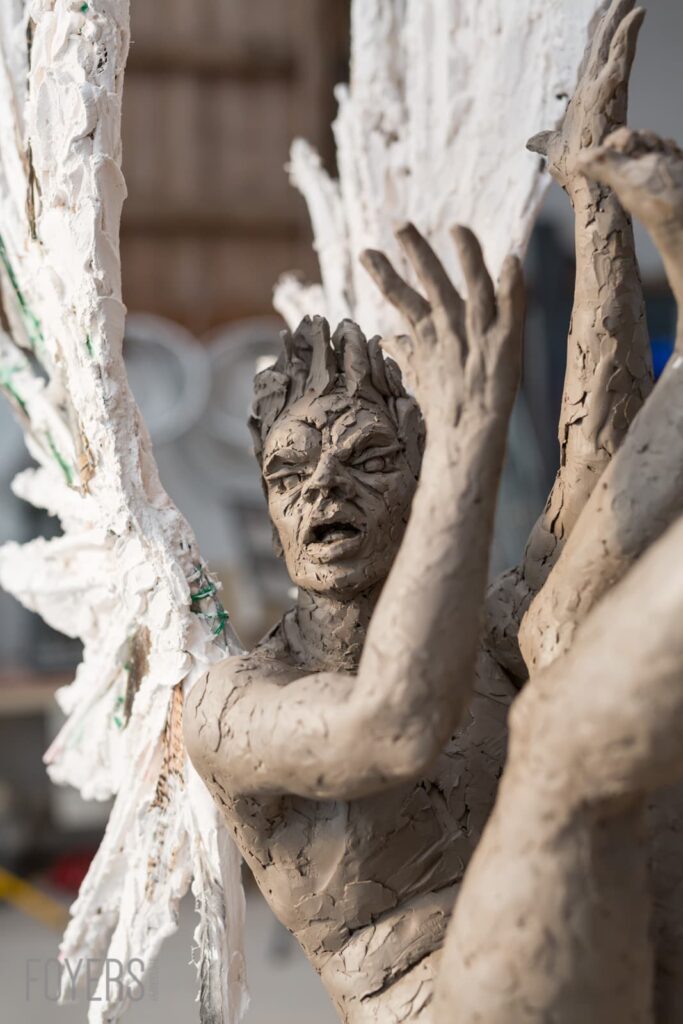 Clay sculpture of a distressed human face and raised arms, partially covered with white plaster or paint.