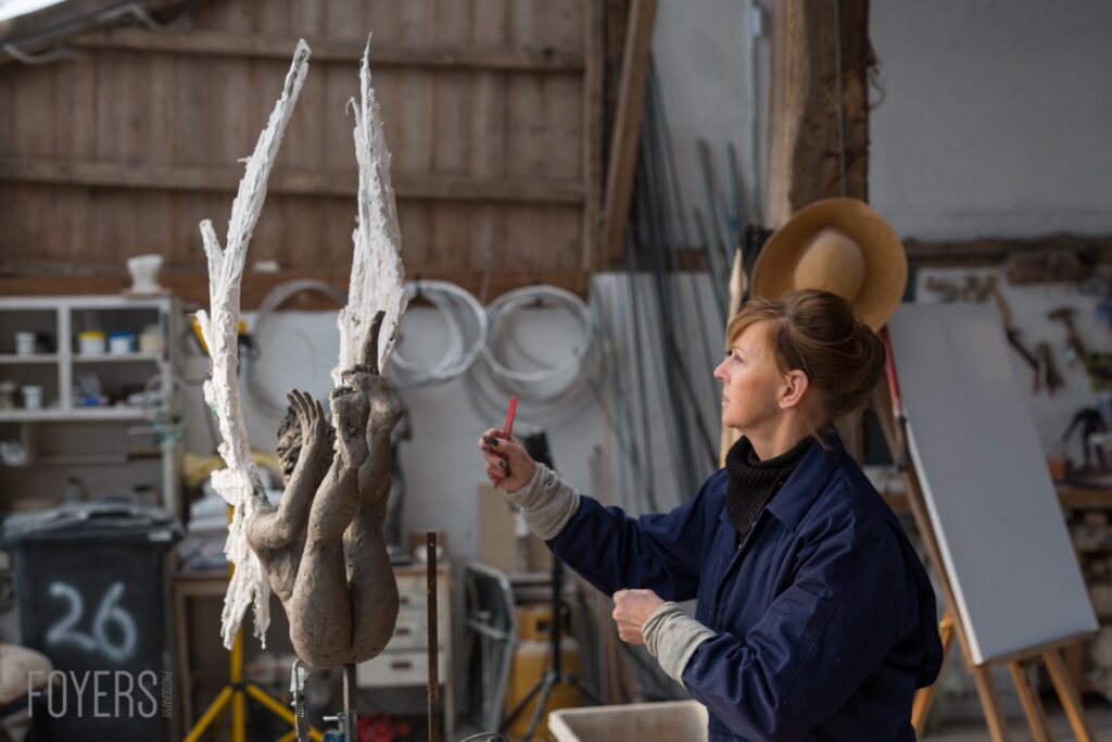 A sculptor works on a clay figure in a studio, applying white plaster to outstretched arms with a brush.