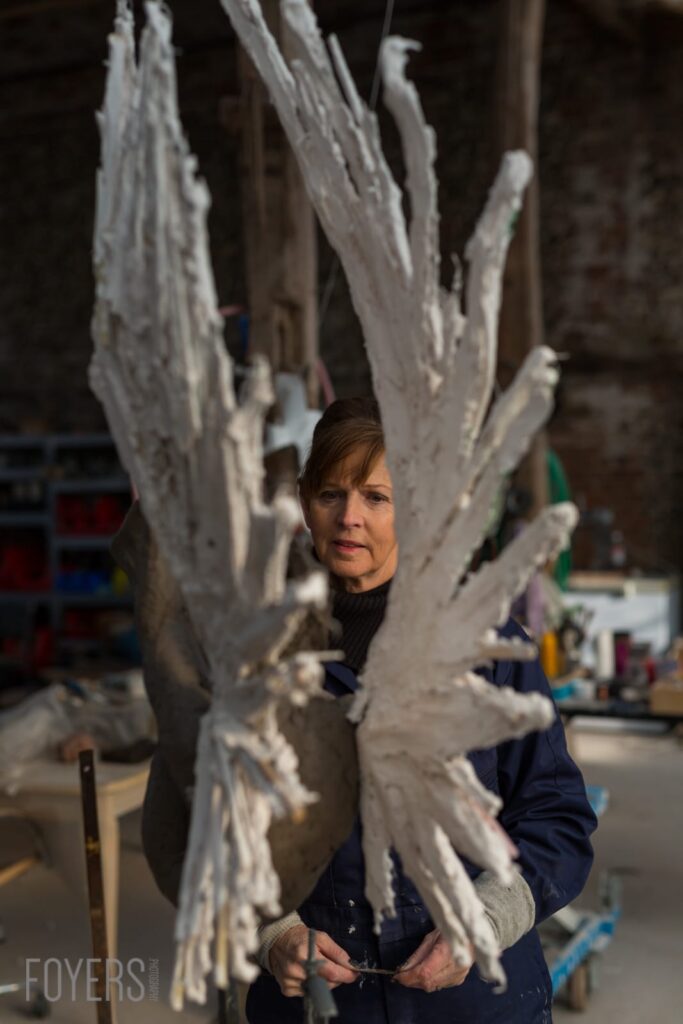 A woman holds a large white plaster wing sculpture in an art studio workshop setting.