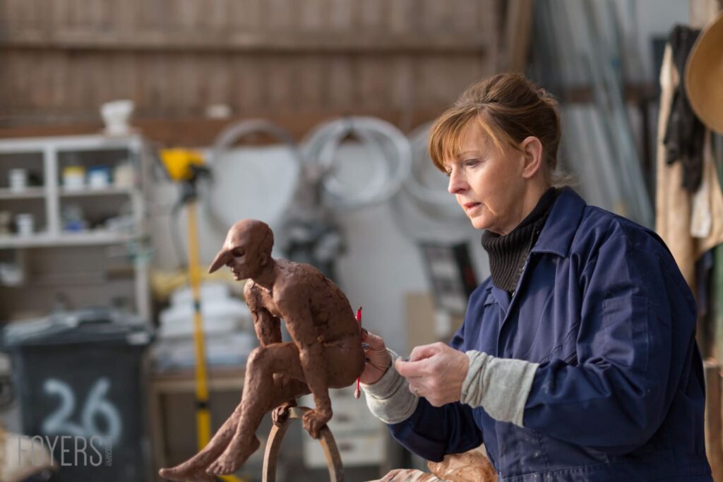 A woman in a blue jacket holds a clay sculpture of a figure in a workshop setting with equipment blurred in the background.