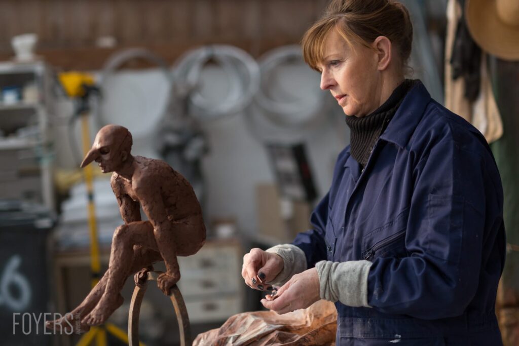 A woman in a blue jacket works on a small wooden sculpture of a seated figure in what appears to be a workshop.