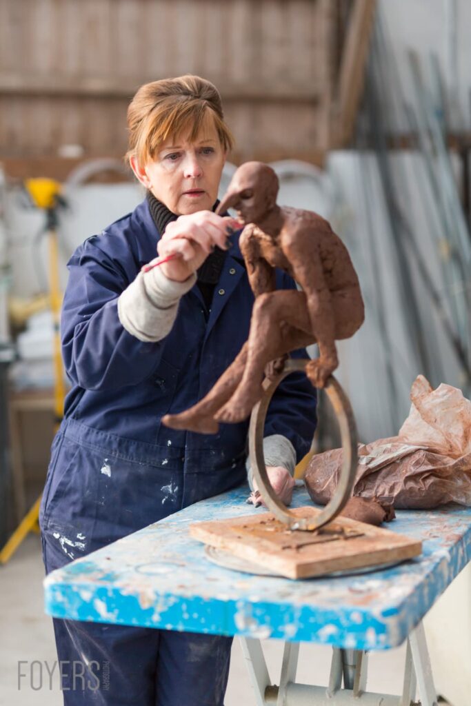 A woman in a navy work coat holds a clay sculpture of a seated figure with a rope, working in a studio.