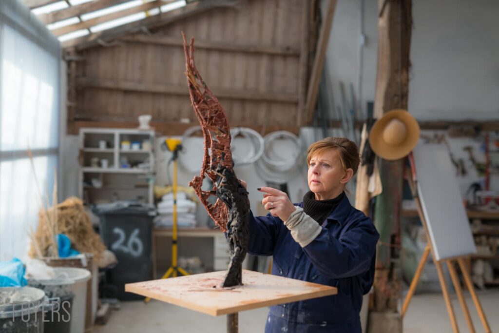 A woman in a studio examines a twisted red and dark sculptural form on a wooden table.