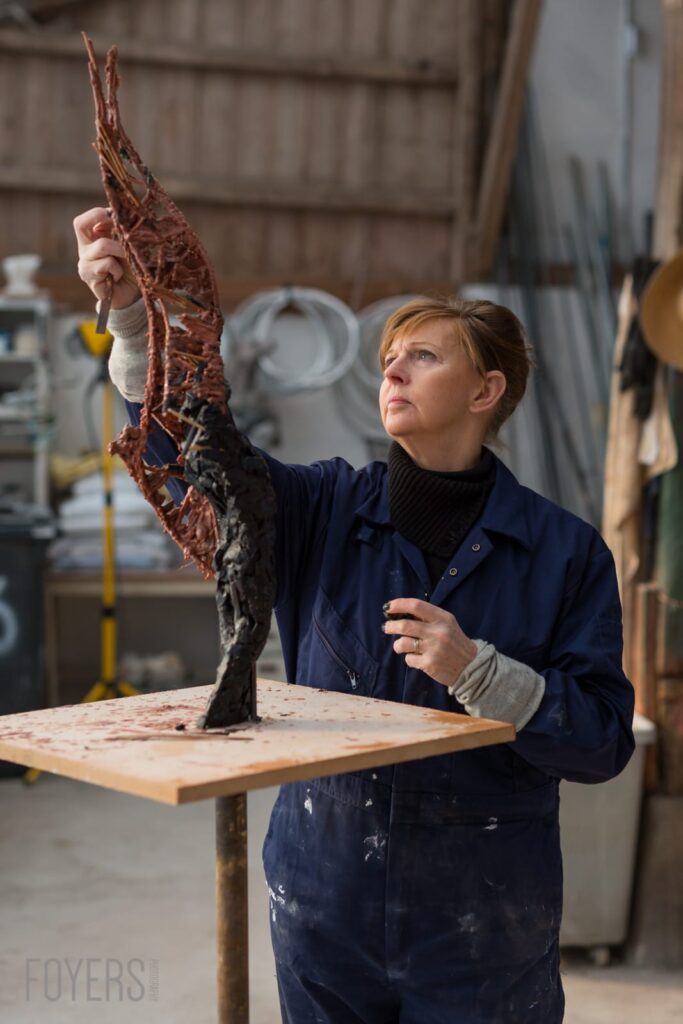 Woman in blue work jacket holding up a red textured sculptural artwork in an industrial studio space.
