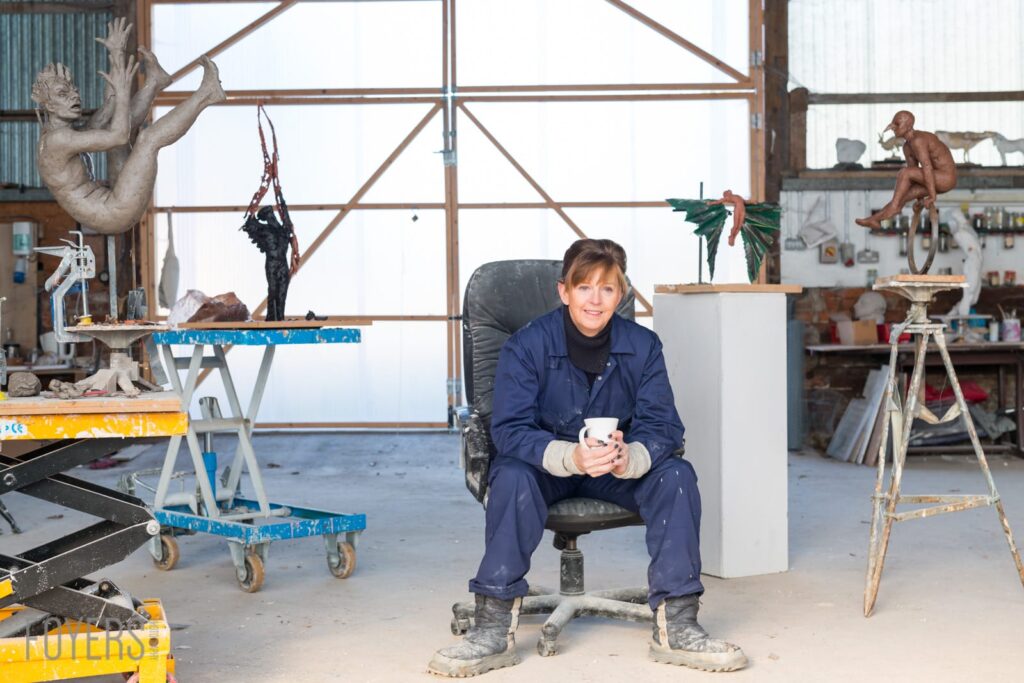 A sculptor sits in a black chair in a studio, holding a cup. Sculptures and tools surround them.