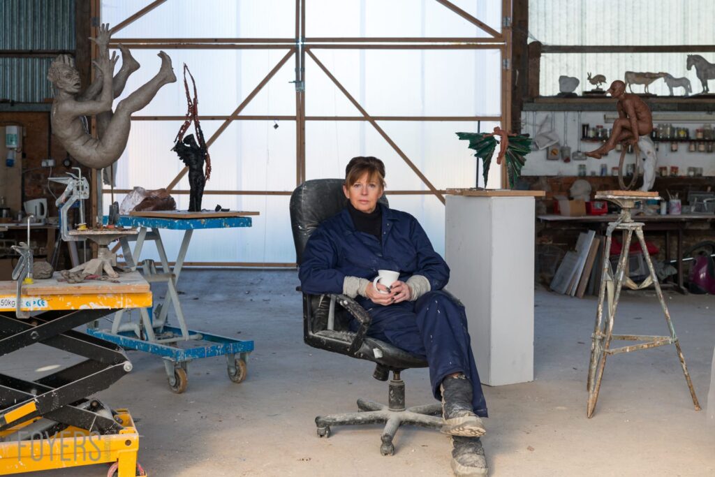 A sculptor sits in a worn chair in a studio workshop, surrounded by various bronze and stone sculptures and tools.