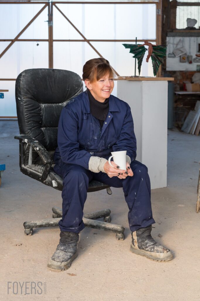 Young boy in navy workwear sitting on a worn office chair in a warehouse, holding a cup and smiling.