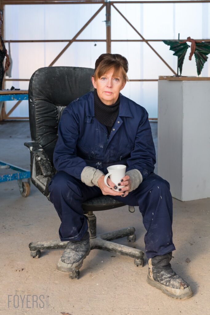 Woman in navy work overalls and grey gloves sits on a worn office chair holding a white cup in an industrial warehouse.