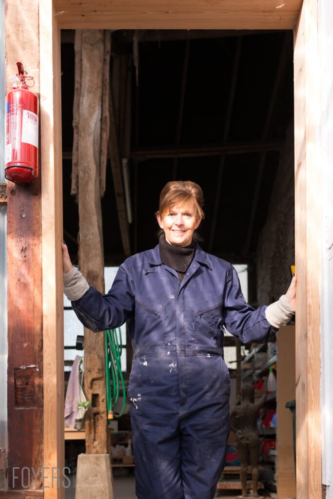 A woman in navy blue overalls stands smiling in a barn doorway, with a fire extinguisher visible on the left wall.