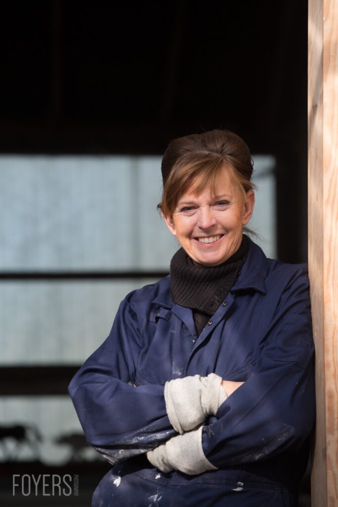 A woman in a navy work jacket and black turtleneck smiles at the camera, arms crossed, leaning against a wooden post.