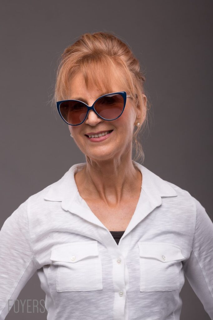 A woman with reddish-blonde hair styled up smiles at the camera, wearing blue-framed sunglasses and a white shirt against a grey background.
