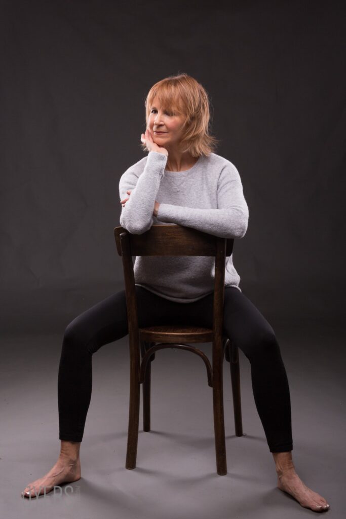 A woman with shoulder-length blonde hair sits on a wooden chair against a dark background, resting her chin on her hand.