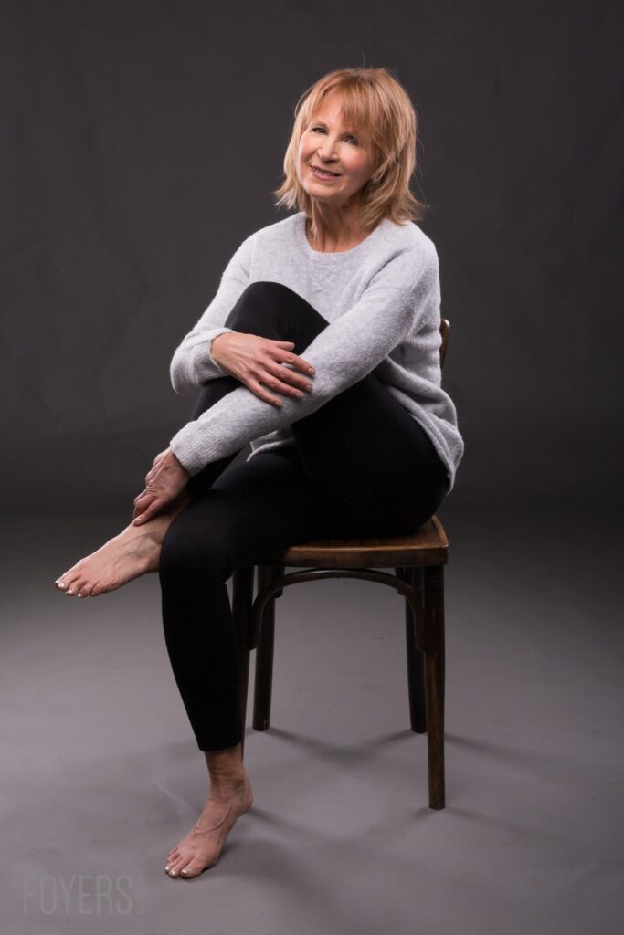 A woman with shoulder-length blonde hair sits on a wooden stool against a dark grey background, smiling at the camera.