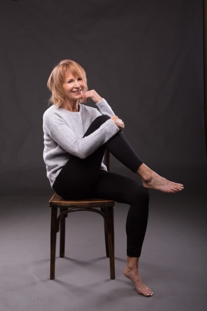 A woman with short blonde hair sits barefoot on a wooden stool against a dark grey background, smiling at the camera.