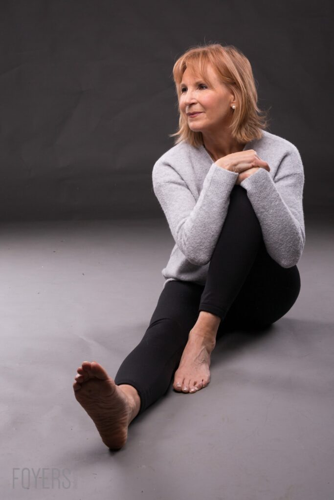 A woman with shoulder-length reddish-blonde hair sits on a grey floor against a dark background, wearing a light grey cardigan and black leggings, barefoot.