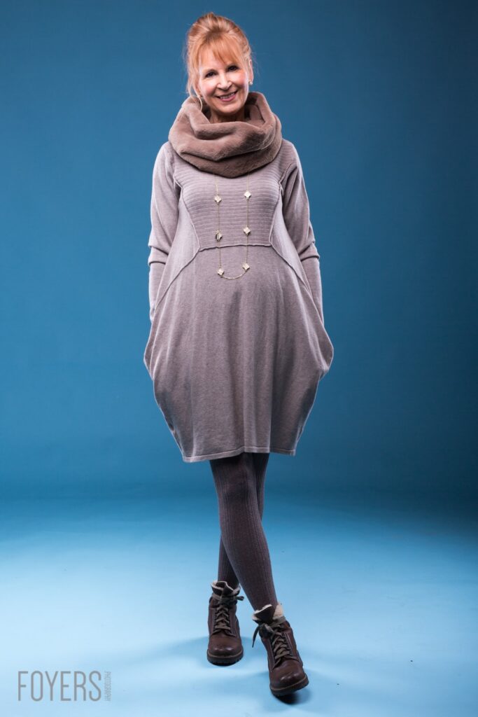 Woman in grey cowl-neck dress with decorative buttons, dark tights and brown lace-up boots, smiling against blue background.