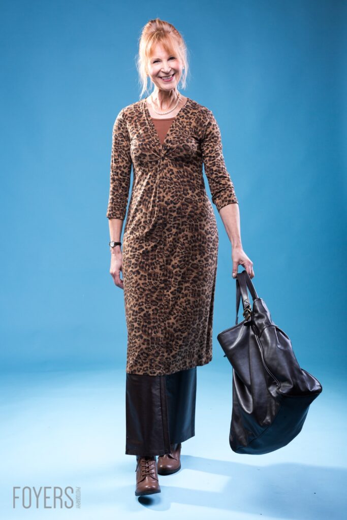 A woman in a leopard print dress and black trousers holds a black leather bag against a blue background.