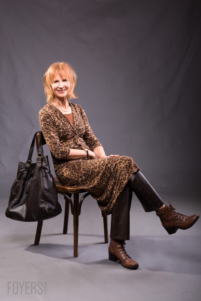 Woman with reddish-blonde hair sitting on a wooden chair, wearing a leopard print dress and brown boots, smiling at the camera with a black bag beside her.