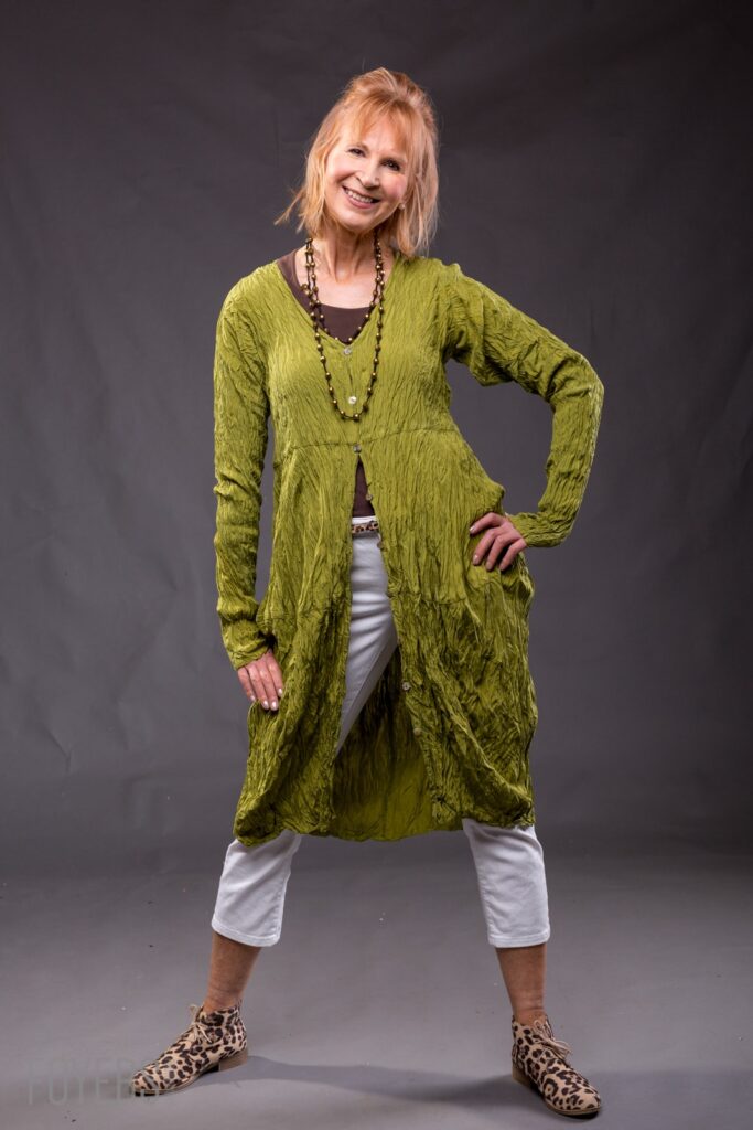 Woman smiling at camera wearing olive green textured cardigan over brown top, white cropped trousers, beaded necklace and leopard print boots against grey background.