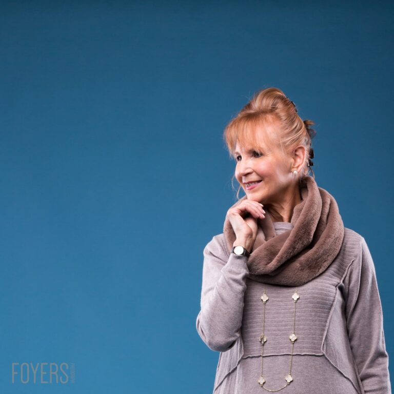 Woman with reddish-blonde hair in a bun, wearing a beige cowl-neck jumper with decorative chains, smiling against a blue background.