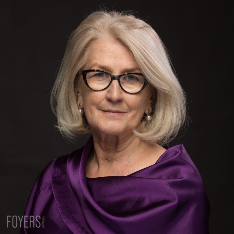 A woman with shoulder-length grey hair and black-framed glasses wearing a purple top, photographed against a dark background.