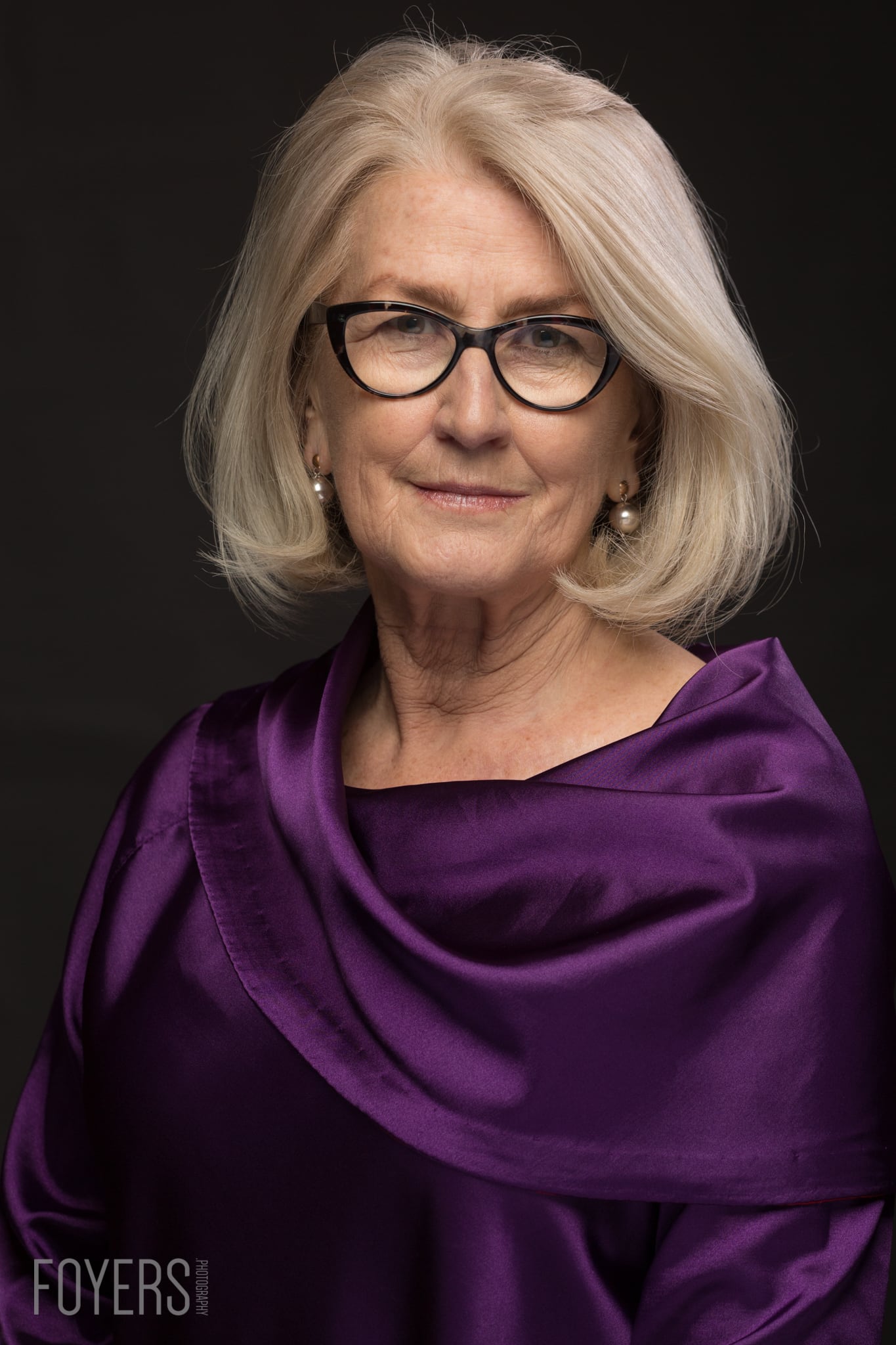 A woman with shoulder-length grey hair and black-framed glasses wears a purple top against a dark background.