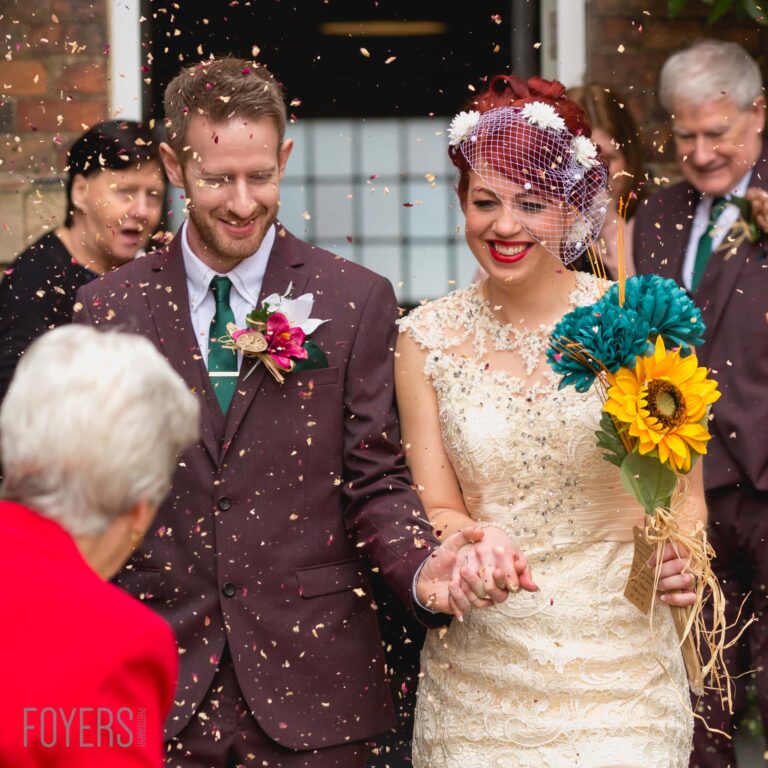 A bride and groom hold hands while confetti falls around them, surrounded by wedding guests smiling at them.