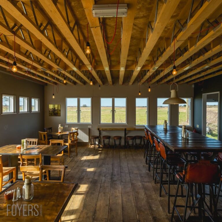 A spacious rustic interior with exposed wooden beams, dining tables, bar seating, large windows overlooking farmland, and pendant lights.