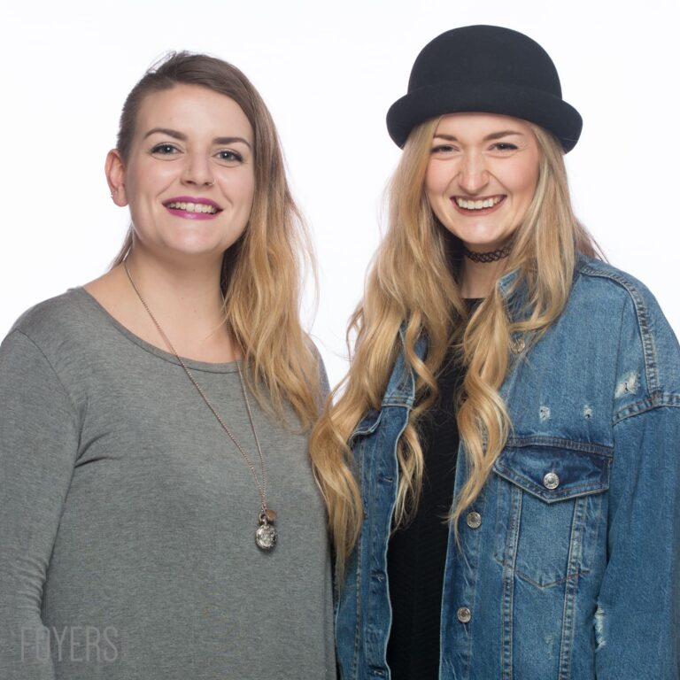 Two young women smiling at the camera against a white background. Left wears grey top with necklace; right wears denim jacket and black hat.