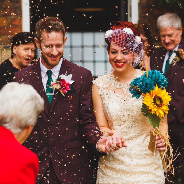 A bride and groom hold hands while confetti falls around them outside a building, surrounded by guests at their wedding celebration.